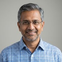 Professional headshot of an Indian structural consultant in his fifties with greying hair, glasses and a checked shirt, photographed against a neutral light grey background.