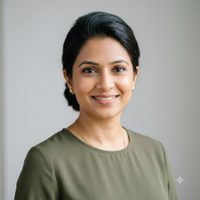 Professional headshot of an Indian interior designer in her thirties with her hair pulled back, wearing an olive blouse, photographed against a neutral light grey background.